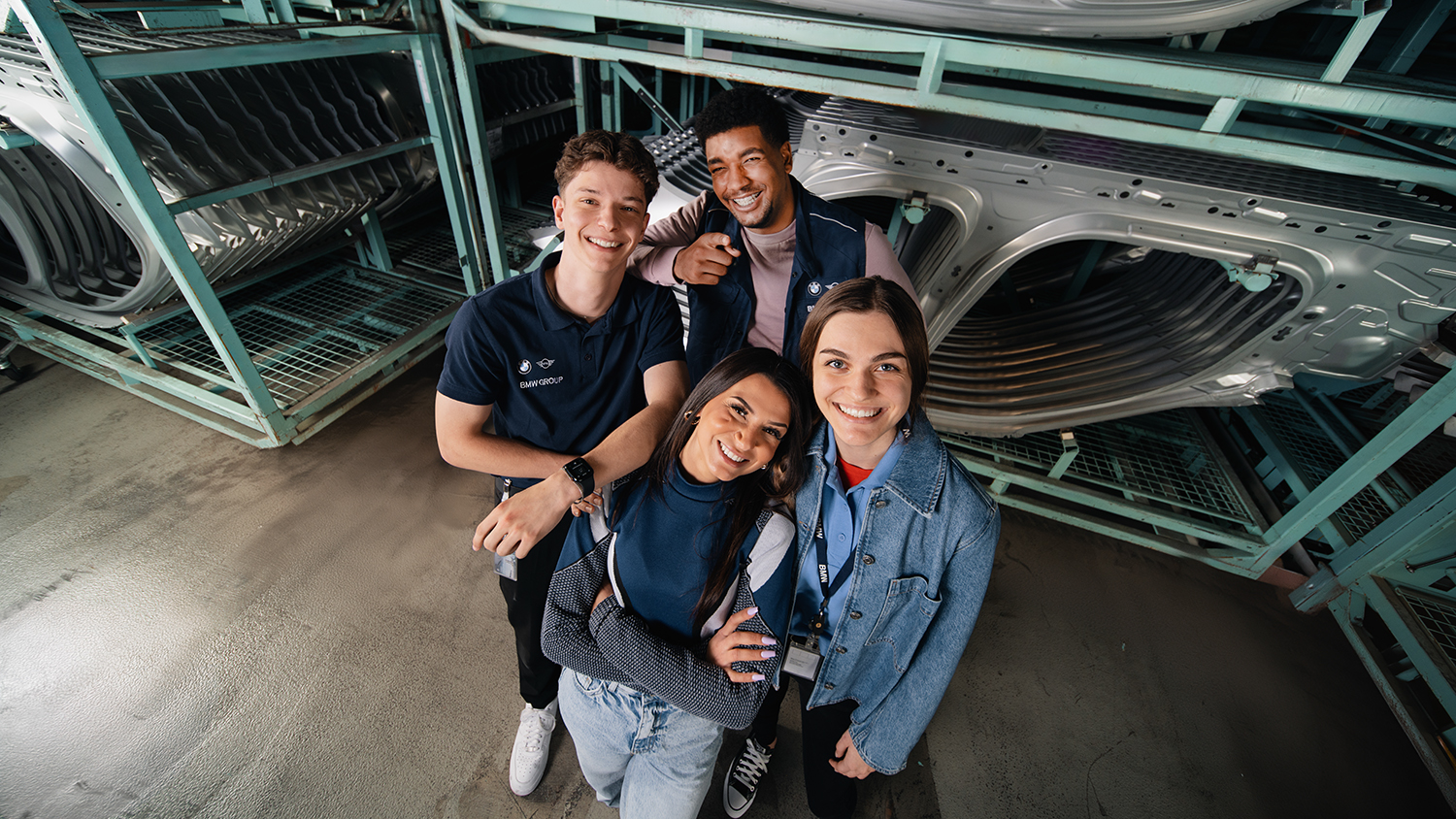A diverse group of four apprentices smiling together in a warehouse setting next to car parts at the BMW Group.