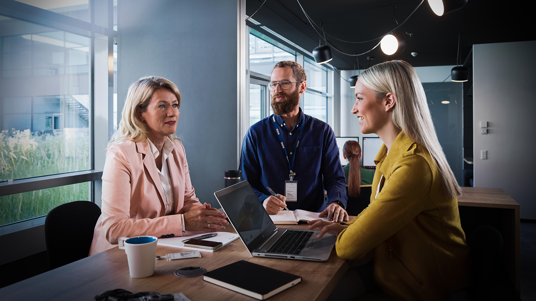 Three colleagues at the BMW Group are having a collaborative conversation in a modern office setting.