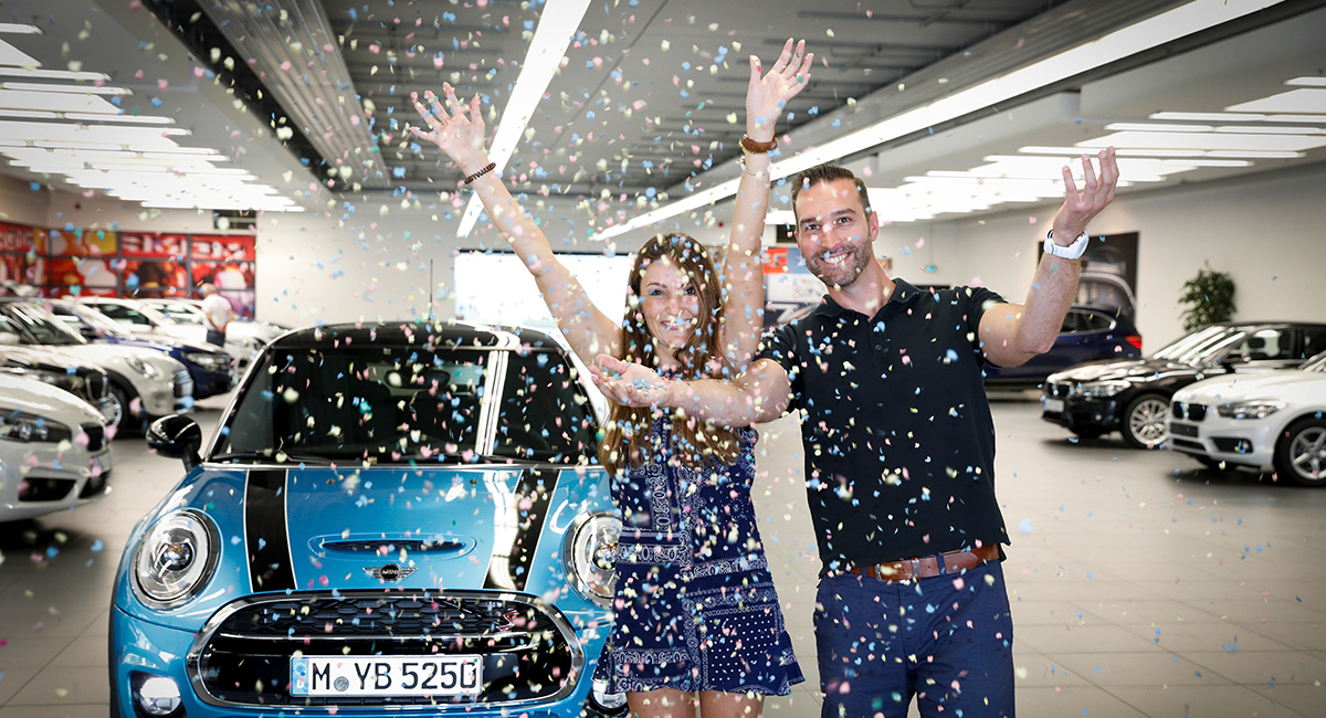 Dieses Bild zeigt eine BMW Mitarbeiterin und einen BMW Mitarbeiter, die sich über ein neues Auto freuen.