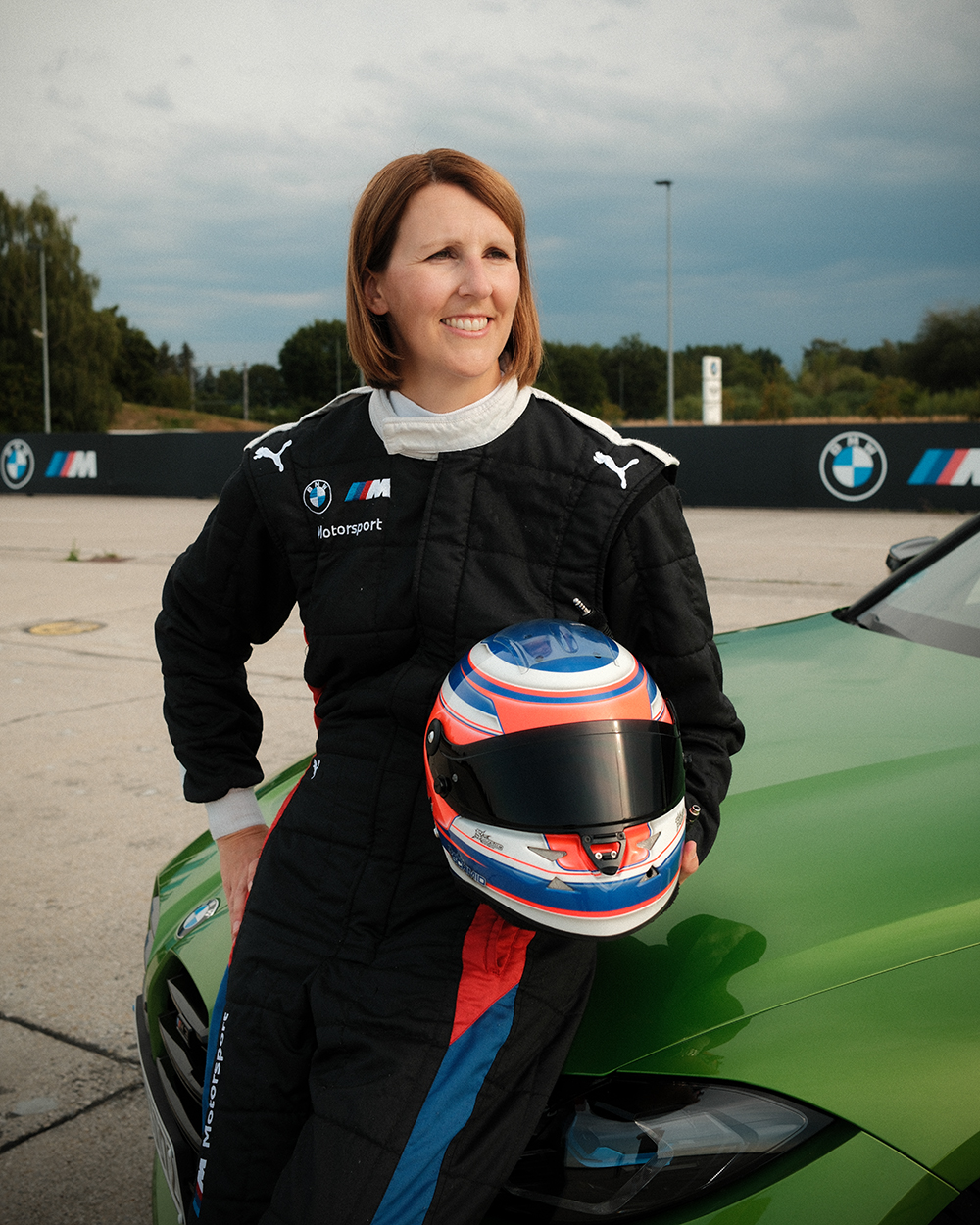 Daniela im Rennanzug mit Helm lehnt an einem grünen BMW M3