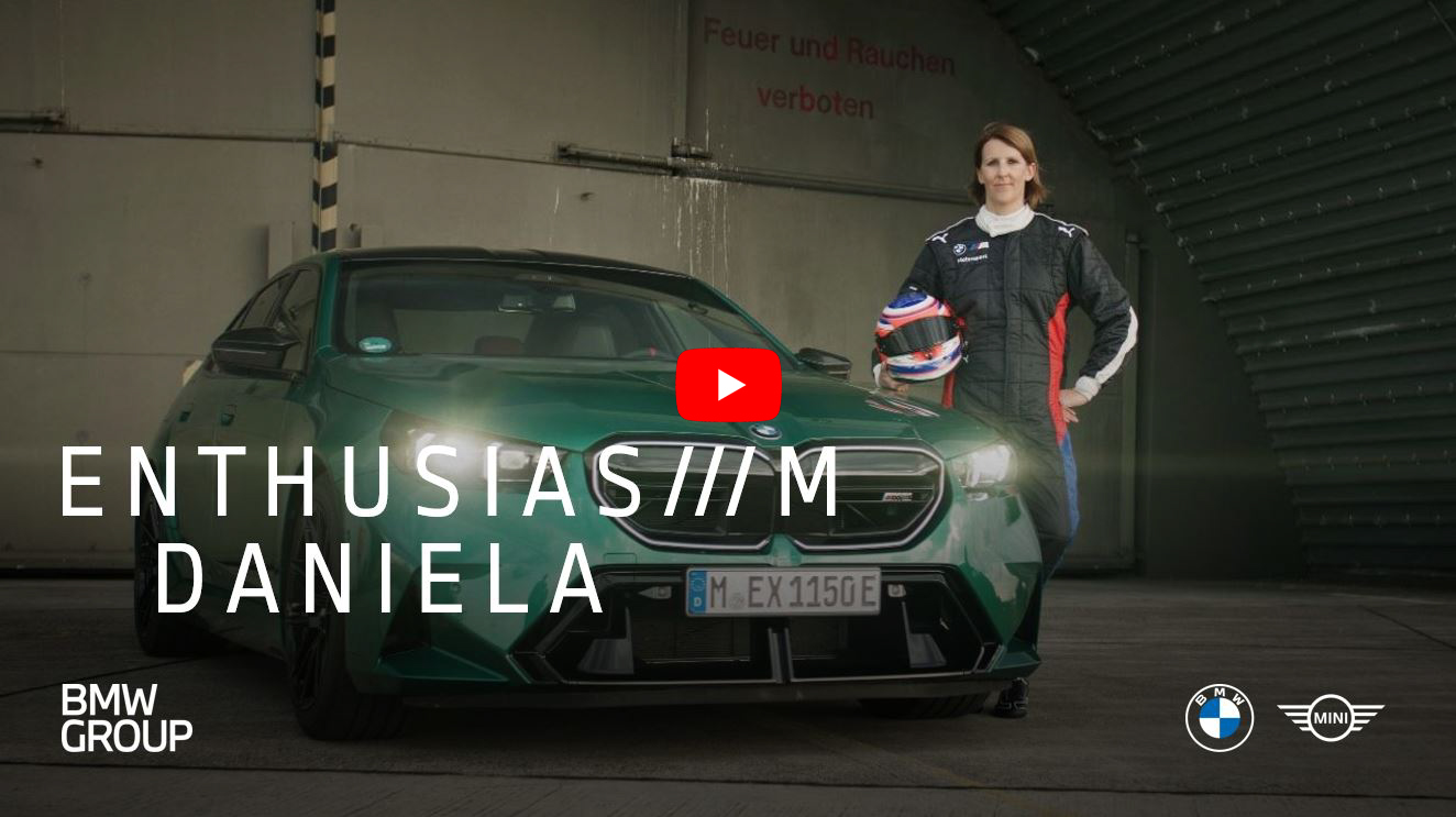 Daniela stands next to a BMW M5 with a racing suit and helmet in her arms