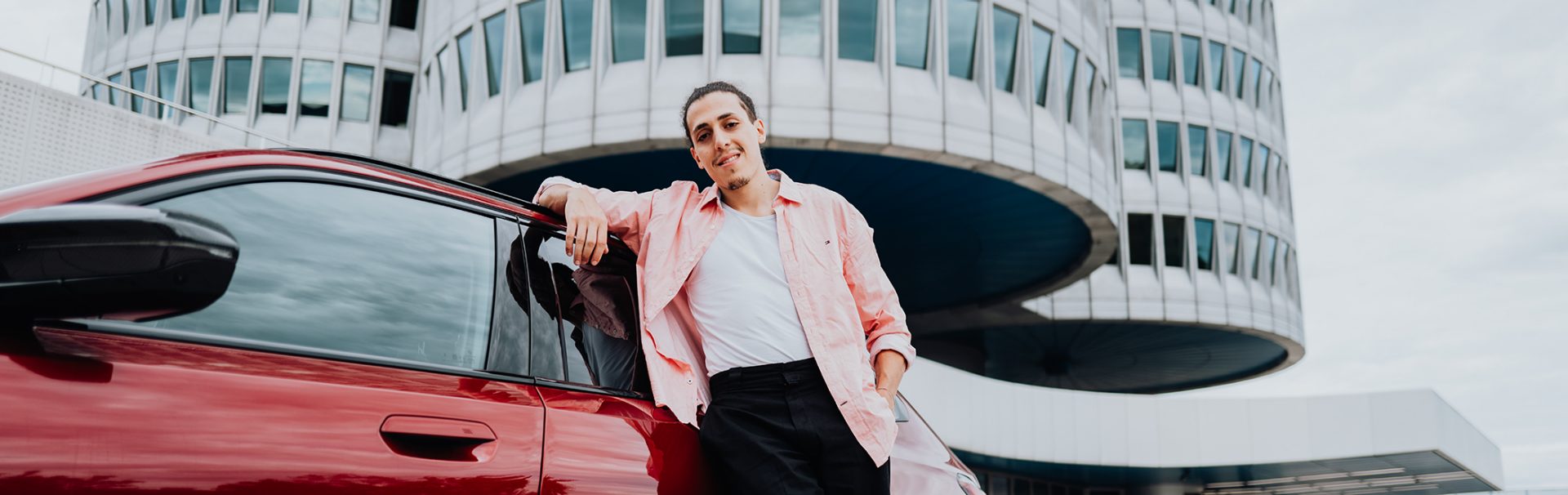 A employee of the BMW Group leaning against a red BMW i5 infront of the BMW four-cylinder.