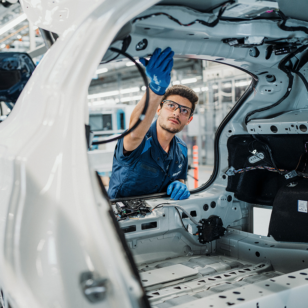The image shows a BMW Group employee working on a car in production.