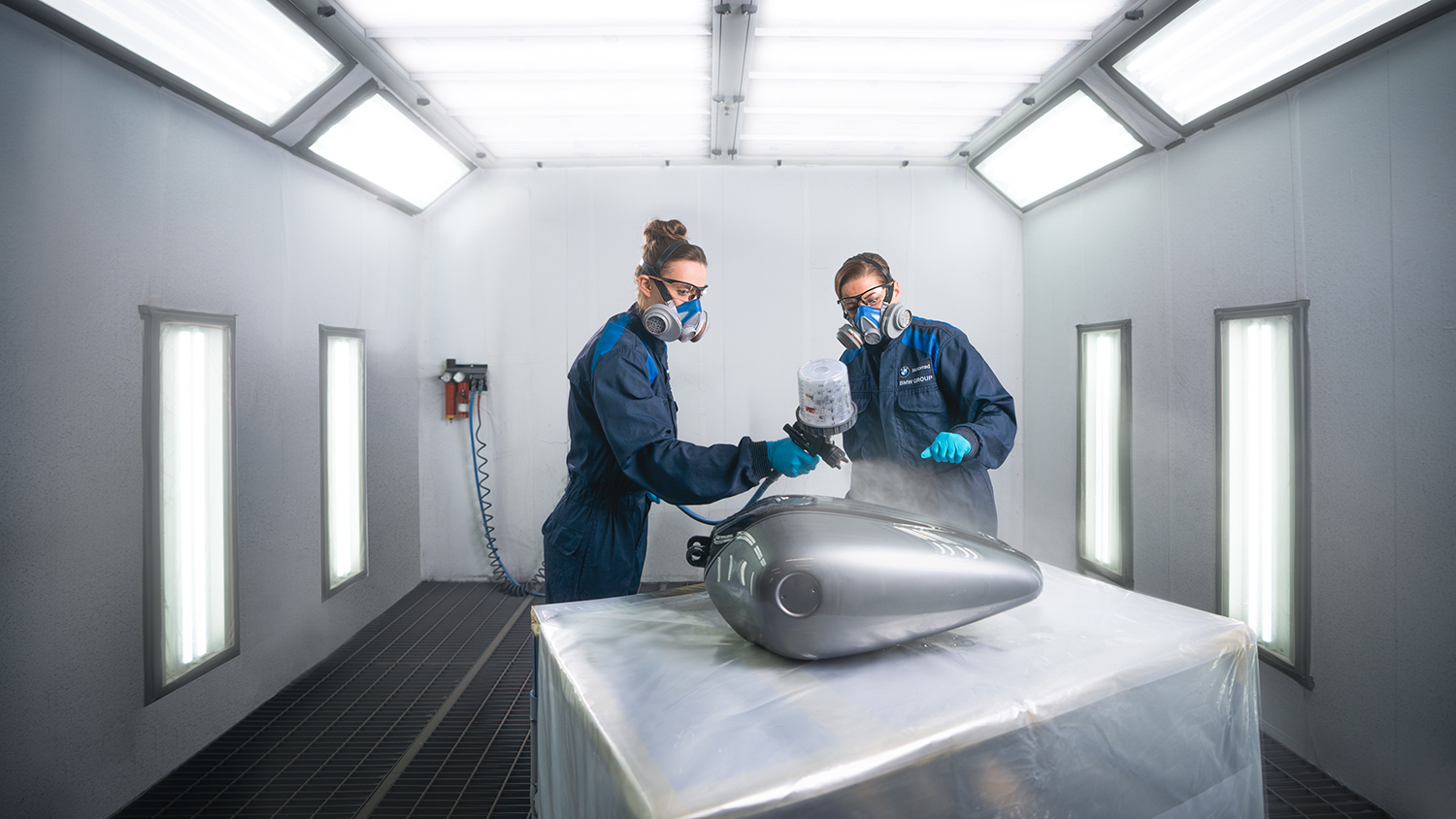 Two employees in the paint shop are working on painting a motorcycle part.