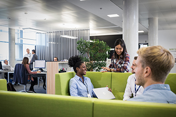 A group of four BMW Group employees sitting together and talking. In the background are colleagues working on their computer.