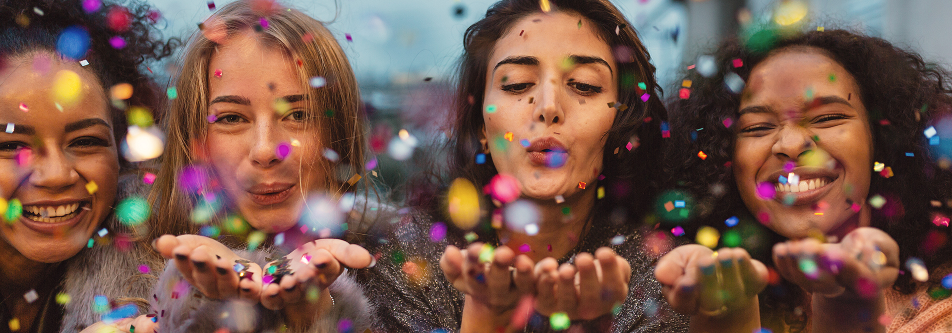 Four women standing next to eachother blowing confetti into the camera.