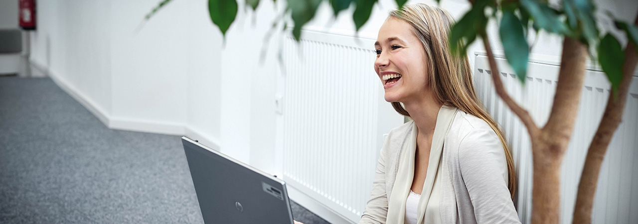 A smiling woman seated beside a laptop, enjoying her work in a bright office environment.
