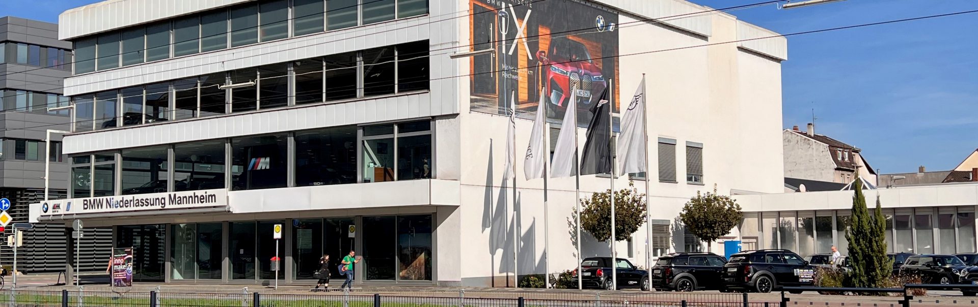 Exterior view of BMW Niederlassung Mannheim dealership with flags and cars.