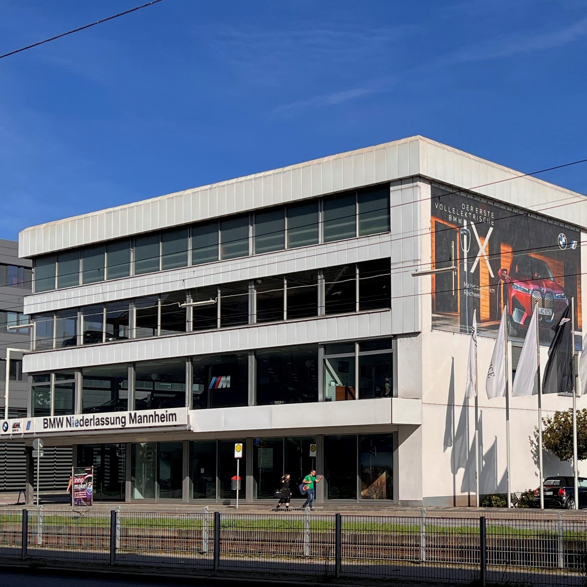 Exterior view of BMW Niederlassung Mannheim dealership with flags and cars.