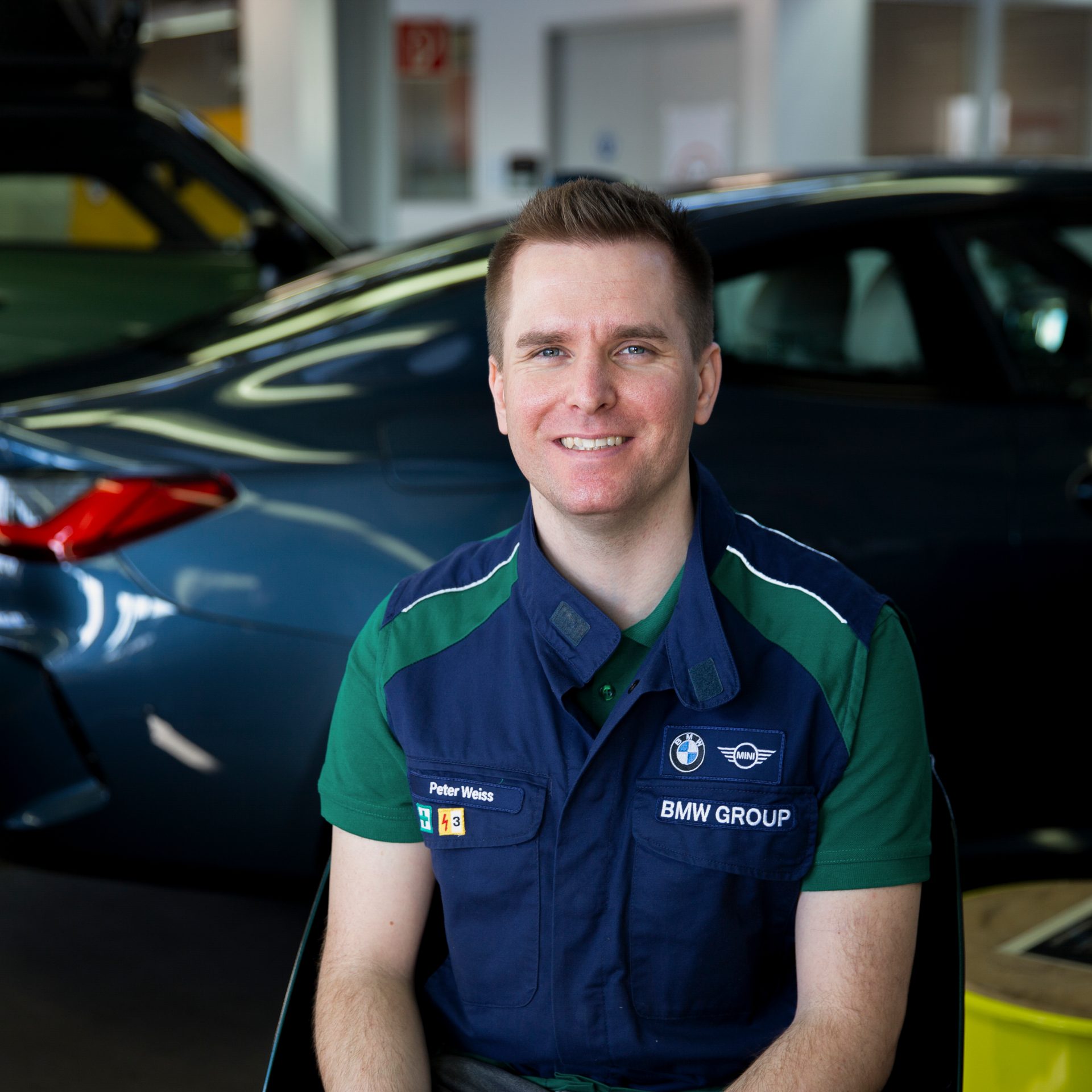 The Trainer for Body and Vehice construction at the BMW Group Plant in Munich sits in front of a BMW and smiles at the camera.