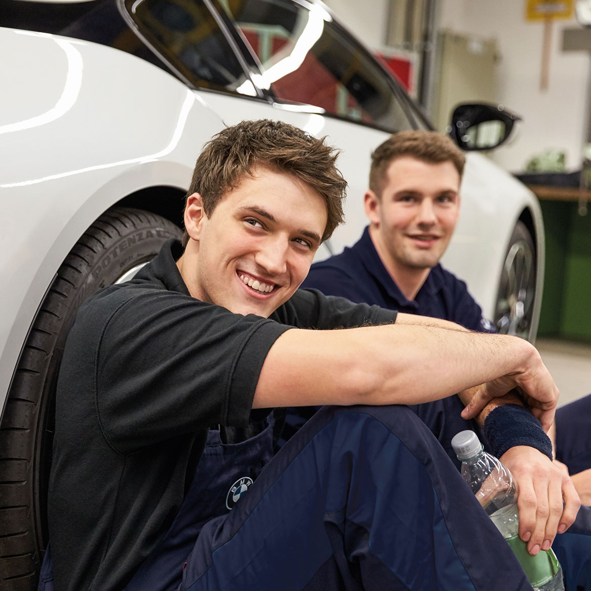 The image shows two apprentices sitting next to a car.