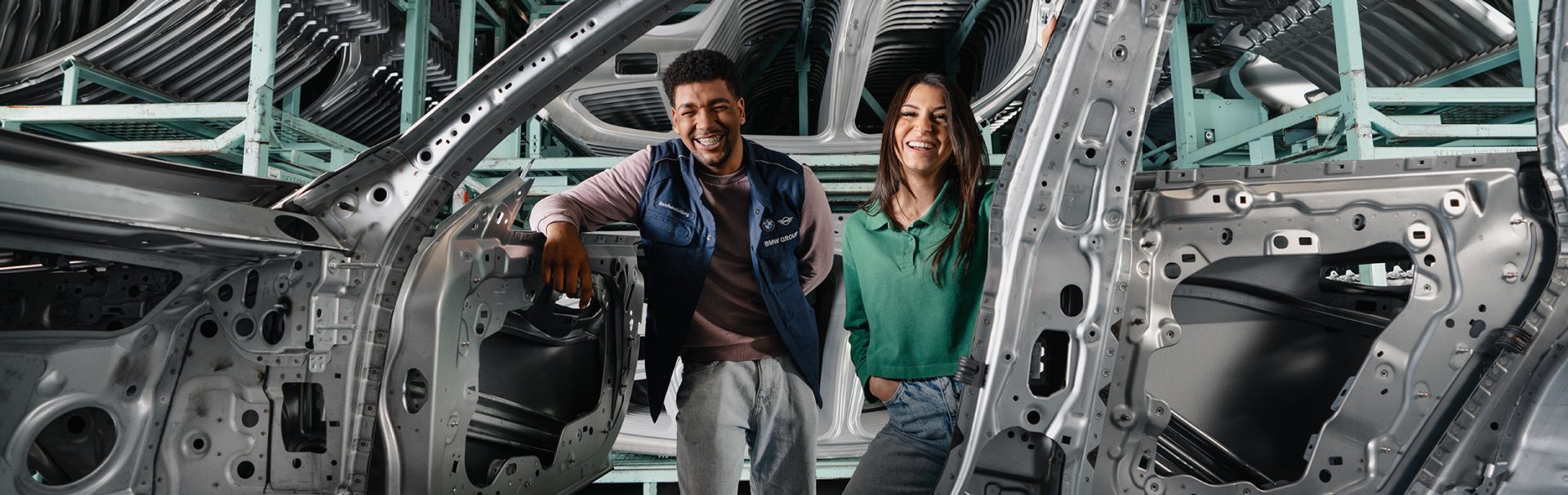 Two BMW Group trainees stand in the production hall and smile through a vehicle body.