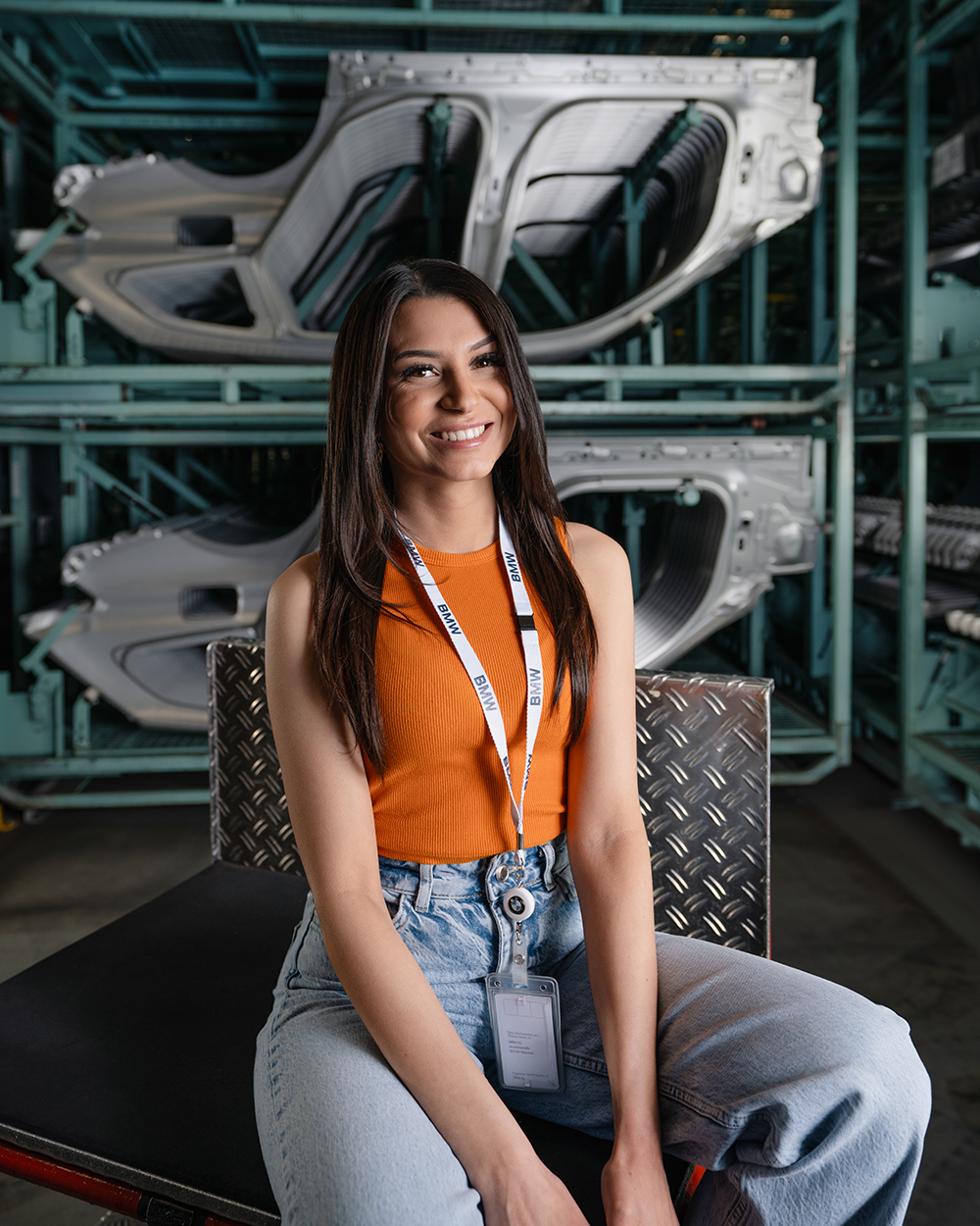 A young BMW apprentice is sitting on a stool and smiles at the camera.