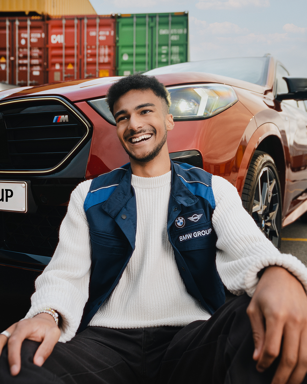 A young BMW apprentice sits in front of a red BMW and is smiling.