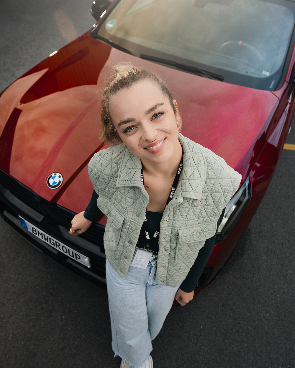 A young apprentice is sitting on the bonnet of a red BMW and smiles at the camera.