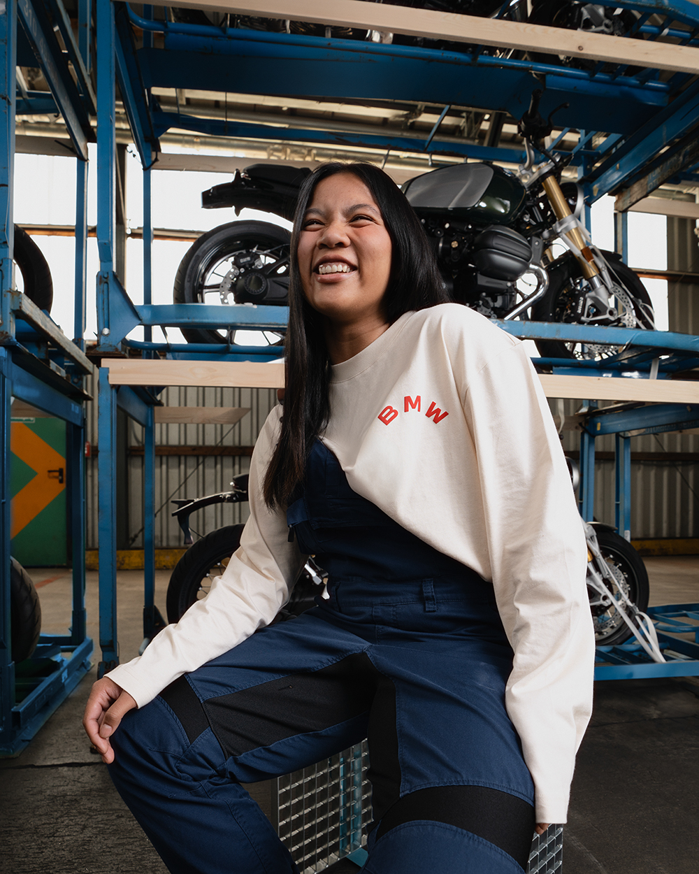 A young BMW apprentice sits on a stool in front of a shelf full with Motorcycles and smiles at the camera.