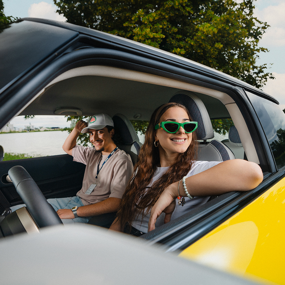  Two cheerful young BMW apprentices enjoying a sunny day in a car, with one wearing sunglasses and the other a cap.