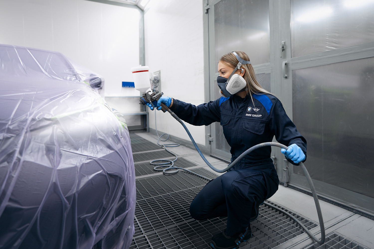 A female apprentice as car sprayer at the BMW Group applying paint to a car using a spray gun in a workshop.