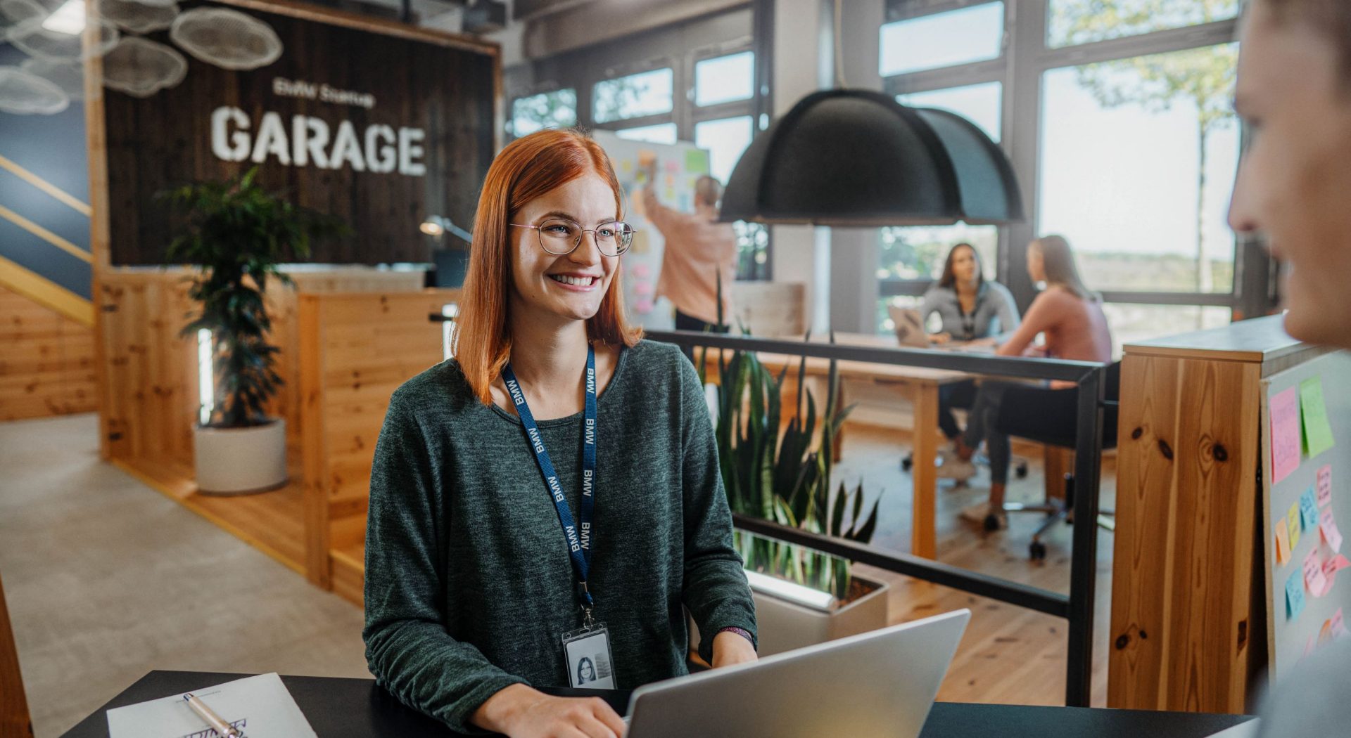 A professional woman with glasses smiling while using a laptop in a modern workspace at the BMW Group.
