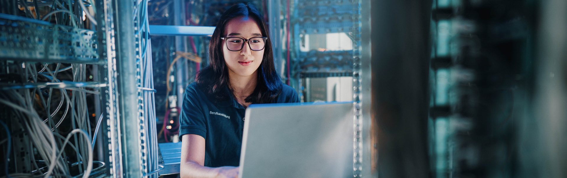 A focused female apprentice using a laptop in a modern data centre filled with networking equipment.
