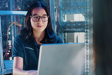 A focused female apprentice using a laptop in a modern data centre filled with networking equipment.