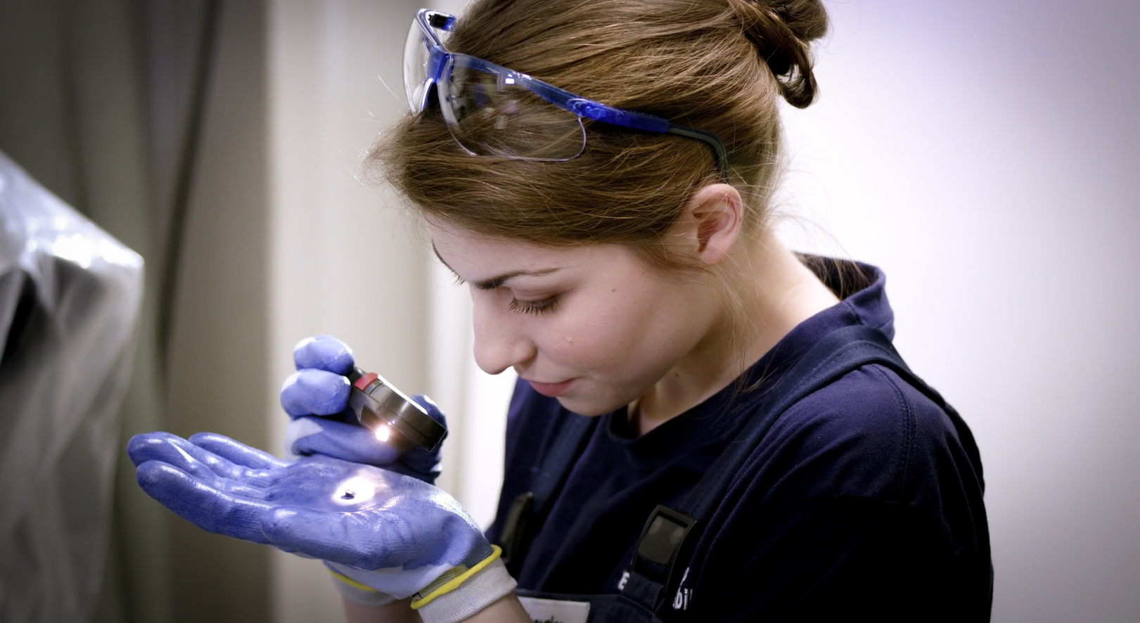 A BMW Group apprentice examines an object on her hand with a magnifying glass.