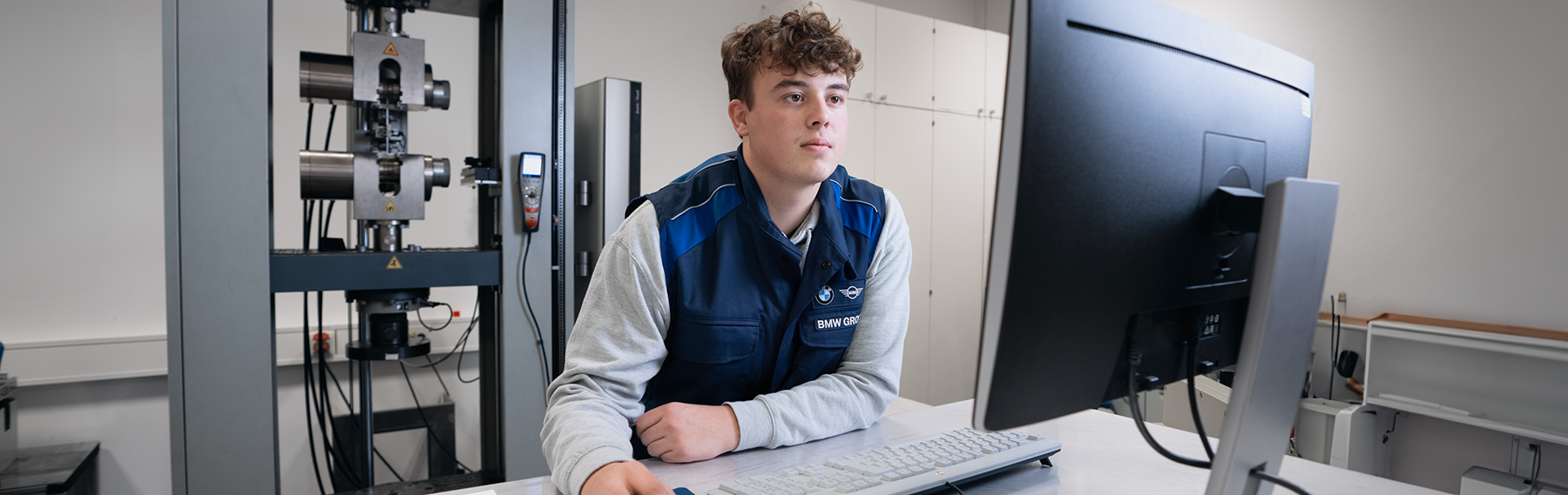 A trainee works at the PC beside a material testing device.