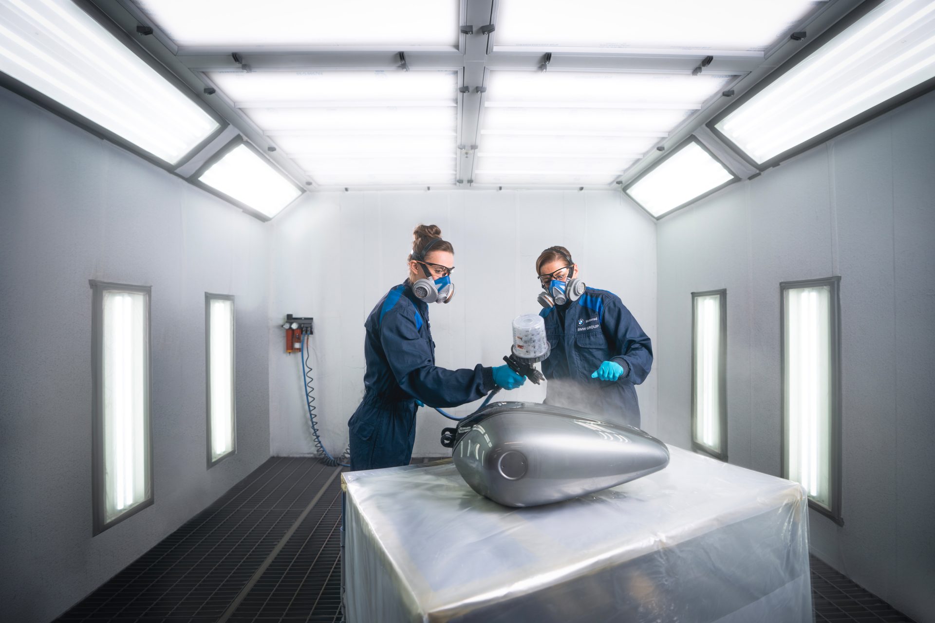 Two process mechanics for coating technology at the BMW Group paint a motorcycle part in a professional paint booth.
