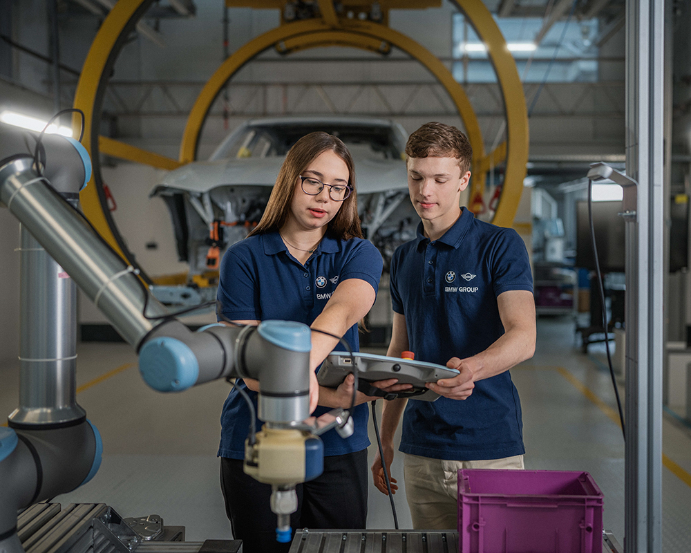 Two dual students having a conversation in the production hall with a BMW M3 in the background