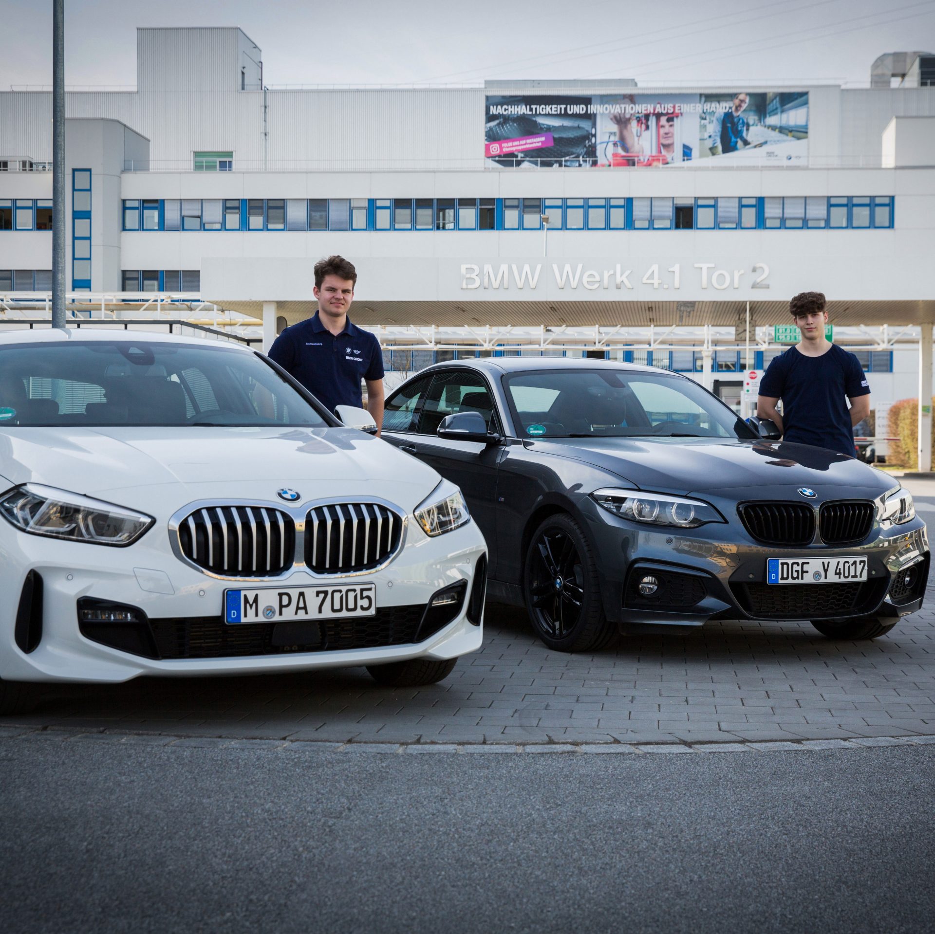 The image shows two apprentices standing next to their cars.