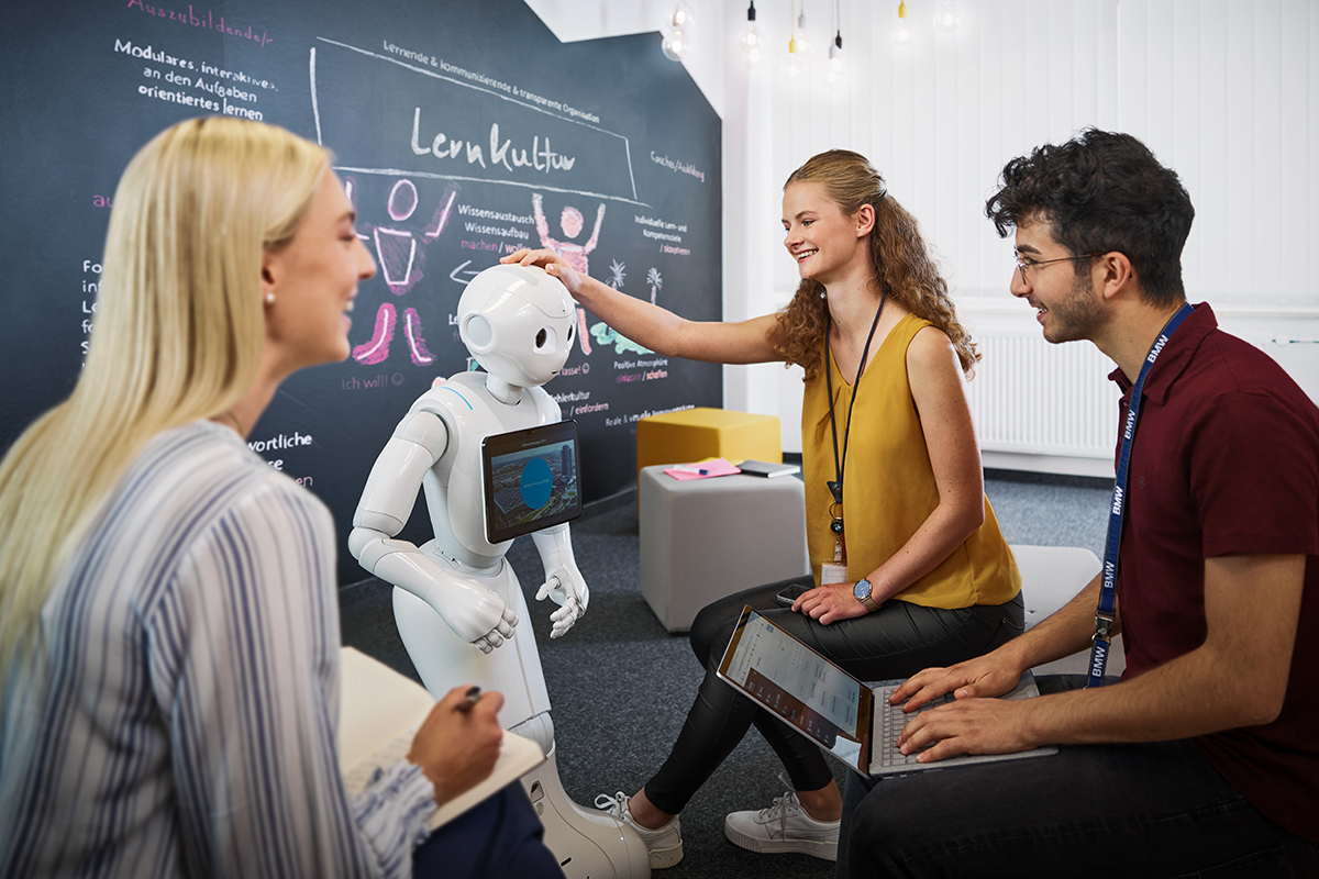 Students for artificial intelligence at the BMW Group interact with a robot in a modern workspace.