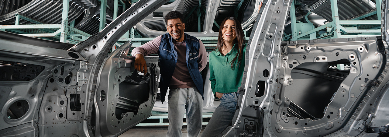 Employees in a BMW Group workshop standing next to a car frame and smile.
