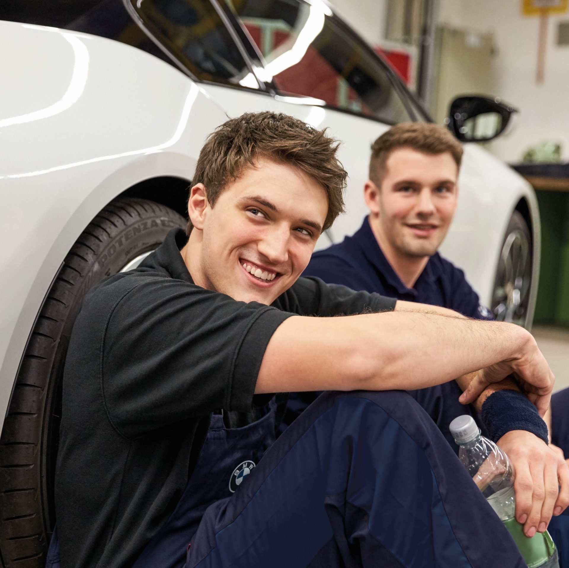 Two apprentices sitting on the floor in a BMW workshop leaning against a car.