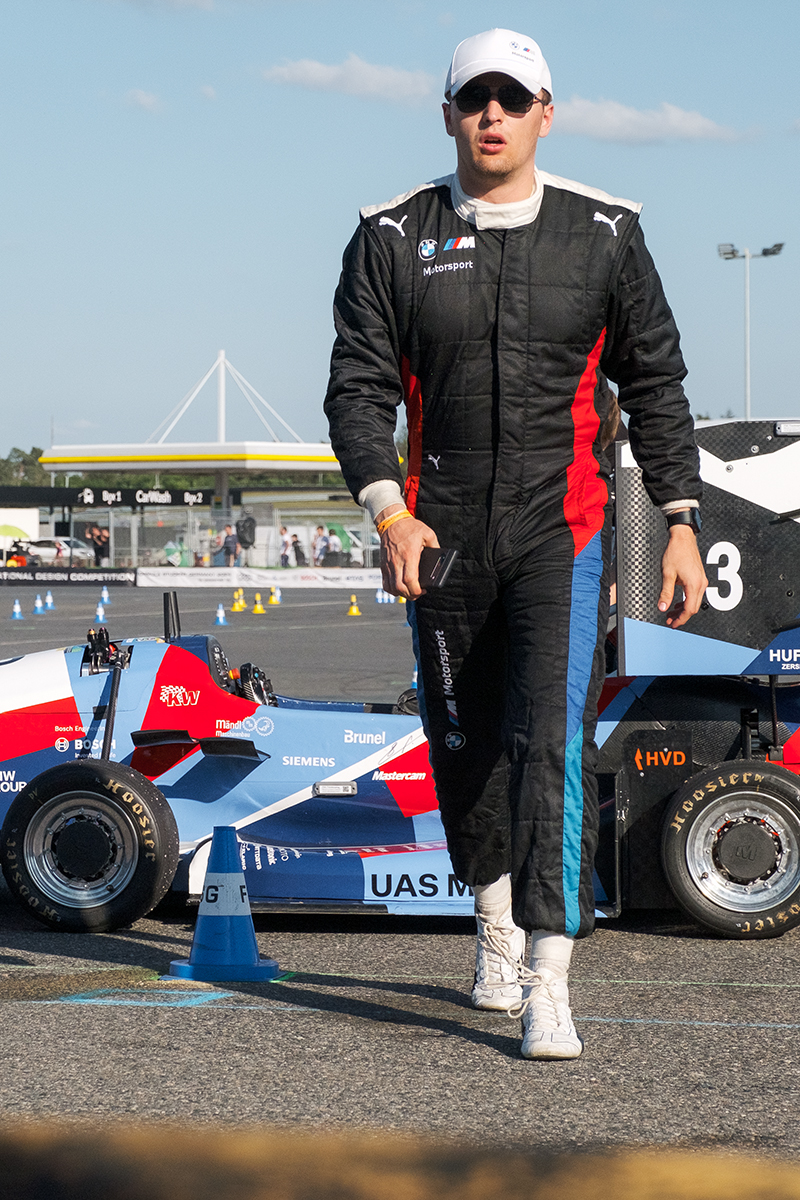 A motorsport driver in a racing suit walking past a race car.
