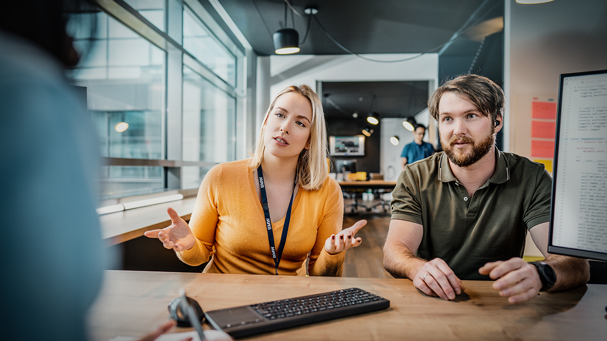 Three colleagues at the BMW Group are having a conversation in a modern office setting.
