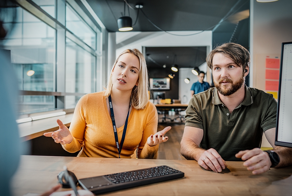Three colleagues at the BMW Group are having a conversation in a modern office setting.