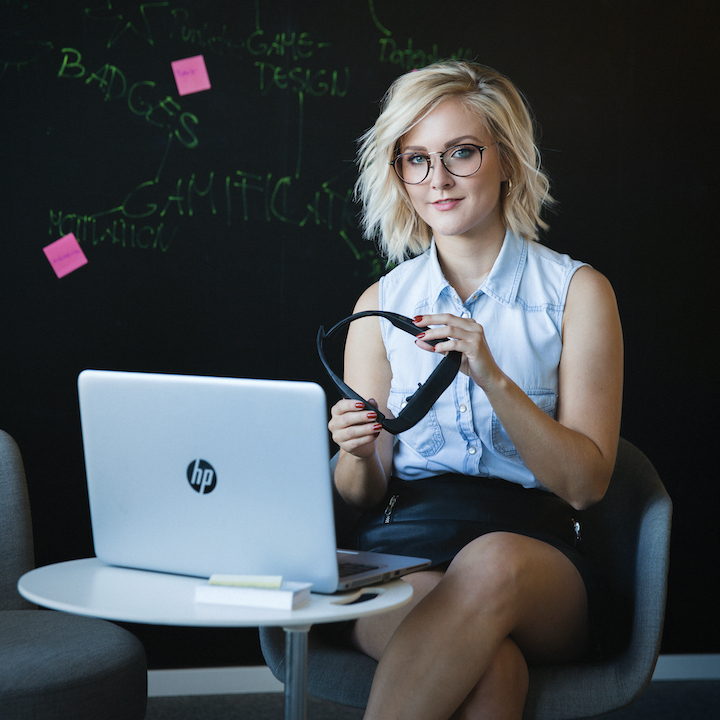 A young woman sitting behind her computer holding a pair of glasses in her hands.