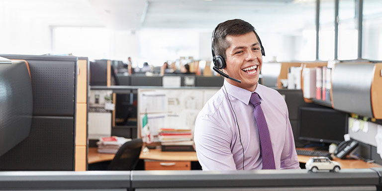 The picture shows a young man, working in customer services, sitting in an office, wearing a headset.