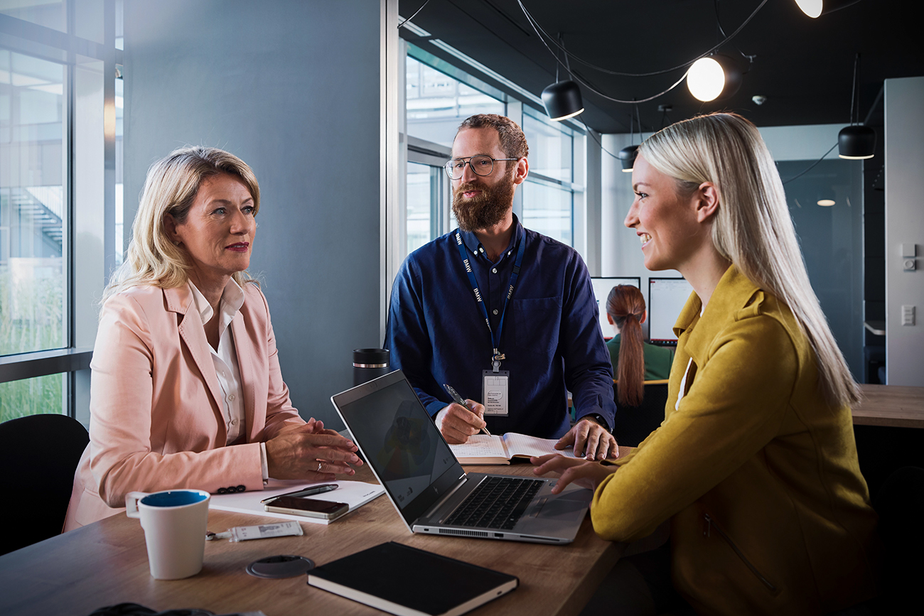 Three colleagues at the BMW Group are having a collaborative conversation in a modern office setting.