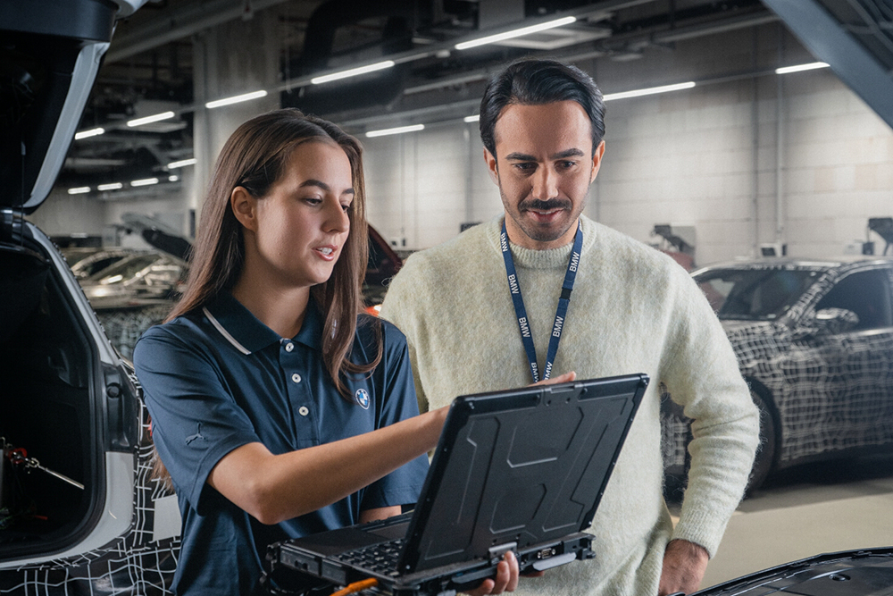 Employees analysing digital information on a laptop beside a vehicle in a workshop setting.