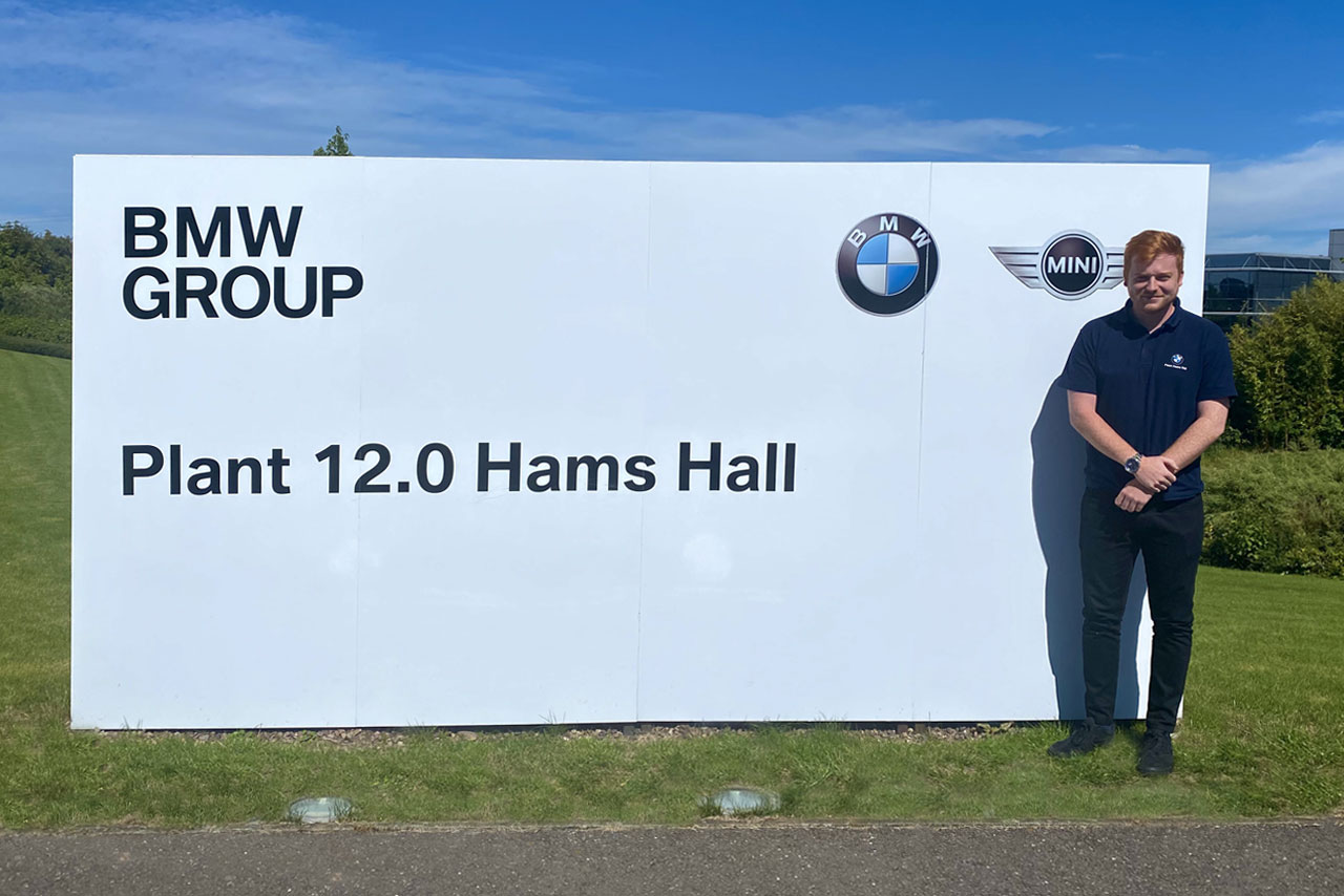 Brad, a Business IT development student standing outside next to a stand-up display with “Plant 12.0 Hams Hall” and the BMW and MINI logos on it