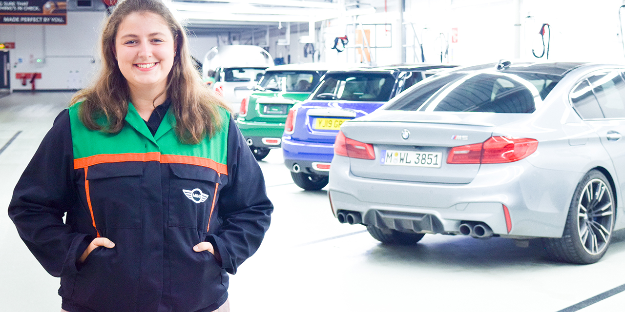 Hannah, an Audit student at the Quality department standing in front of different BMW and MINI cars, smiling at the camera