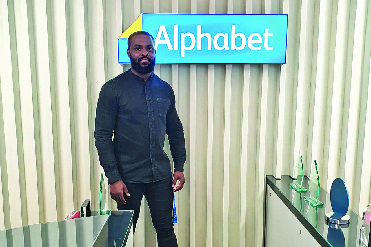 Tobi, a former quality intern now working as a pricing analyst standing in front of a wall with the Alphabet logo at the Alphabet office in Farnborough.