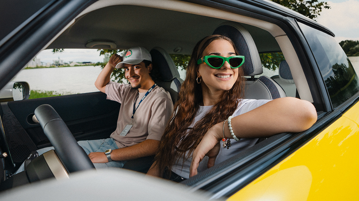 Two cheerful young BMW apprentices enjoying a sunny day in a car, with one wearing sunglasses and the other a cap.
