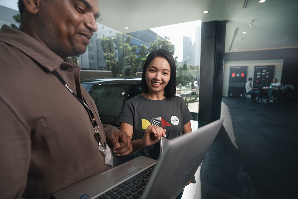 The image shows a BMW Group employee working in purchasing analysing a project with a colleague.