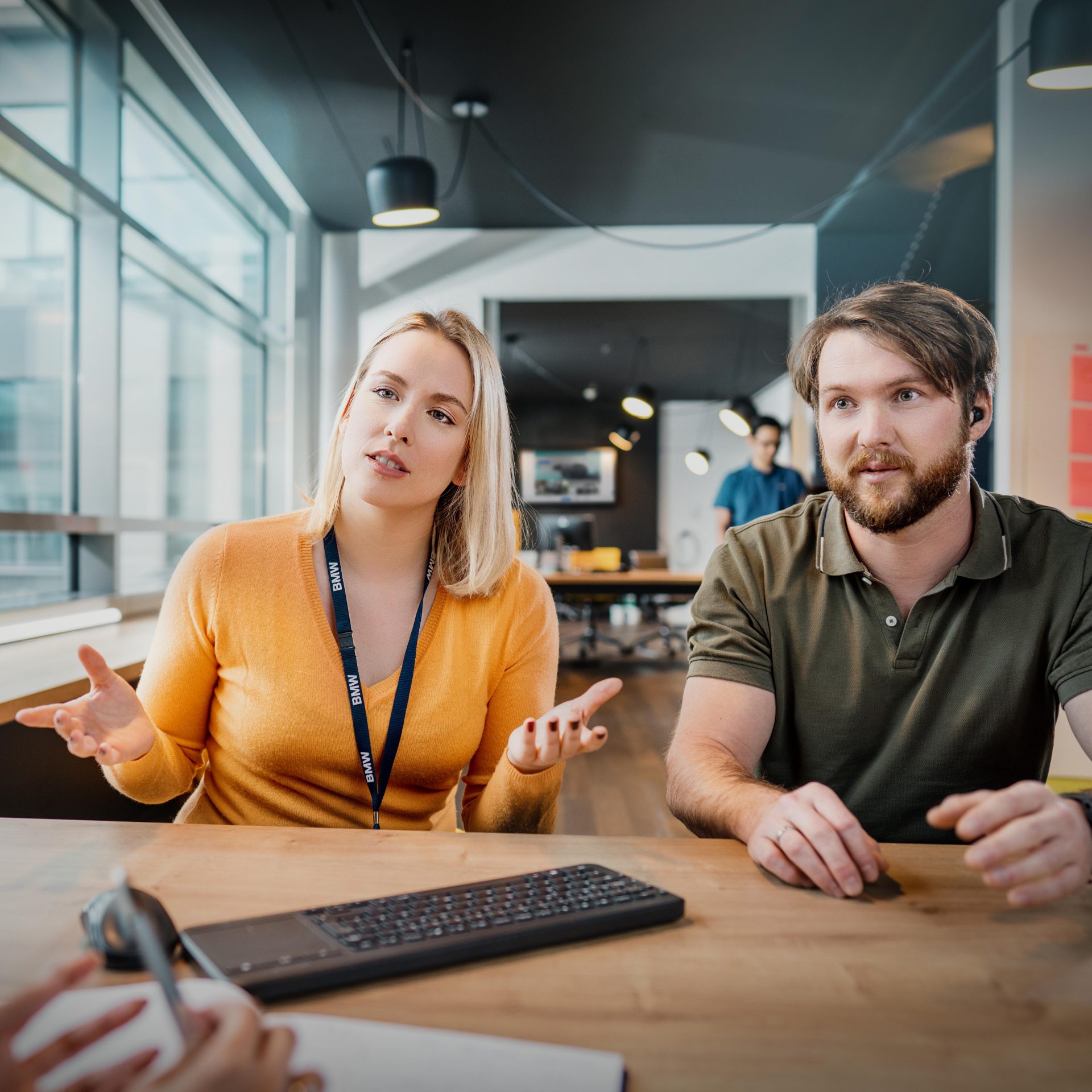 The image shows two BMW Group colleagues at a desk discussing a project.
