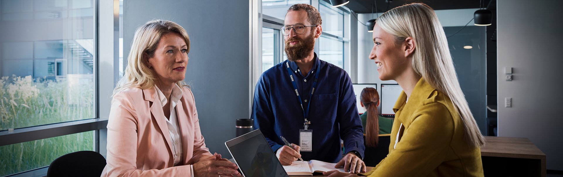 Three colleagues at the BMW Group are having a conversation in a modern office setting.