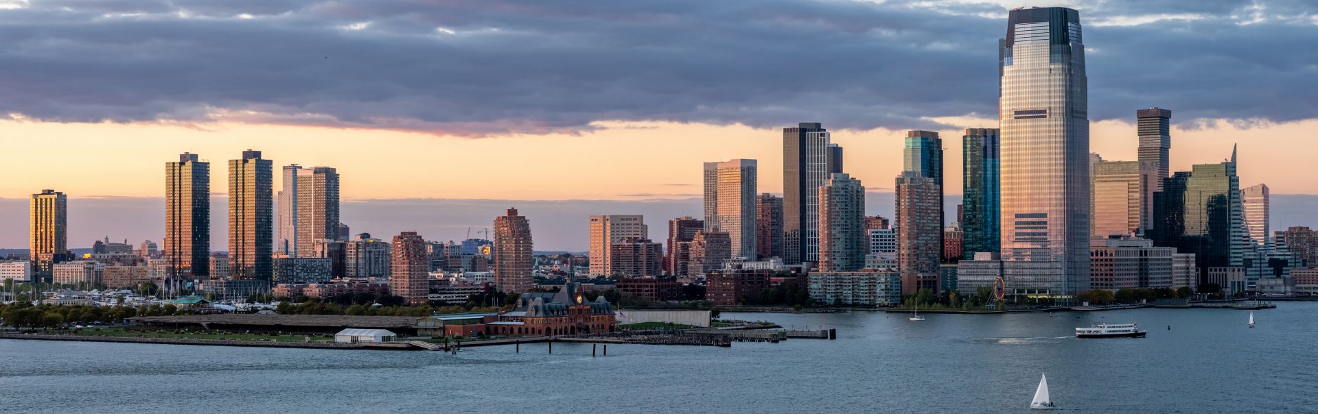 Idyllic landscape showing Jersey City and Hudson River (USA). The image is illuminated by the setting sun. A beautiful cloudy sky with warm summer colours. In the foreground, on the Hudson River, we can see boats and sailboats. The bottom of the sky is fire-coloured.