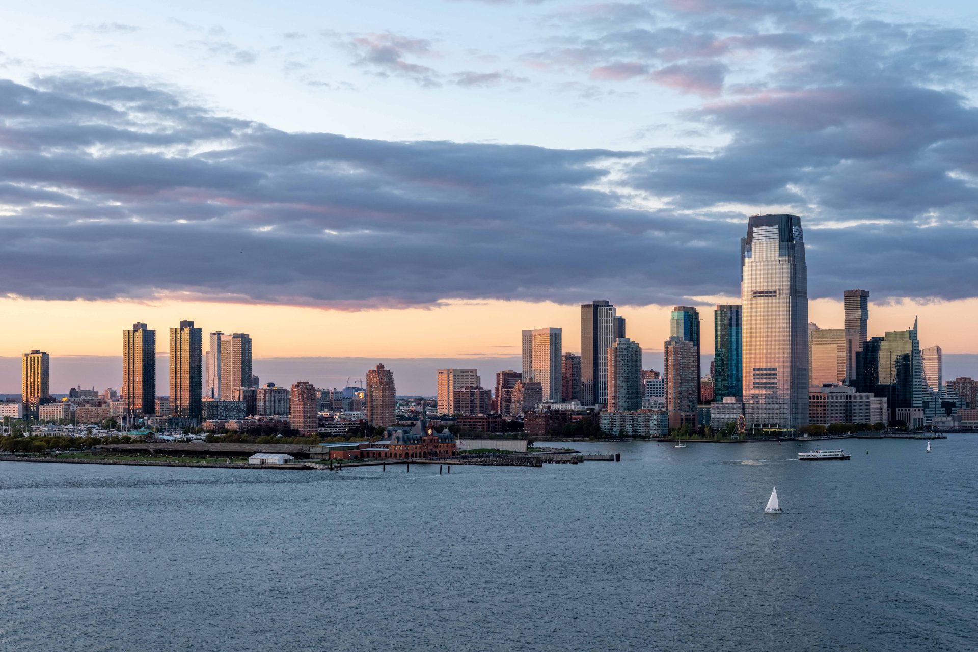 Idyllic landscape showing Jersey City and Hudson River (USA). The image is illuminated by the setting sun. A beautiful cloudy sky with warm summer colours. In the foreground, on the Hudson River, we can see boats and sailboats. The bottom of the sky is fire-coloured.